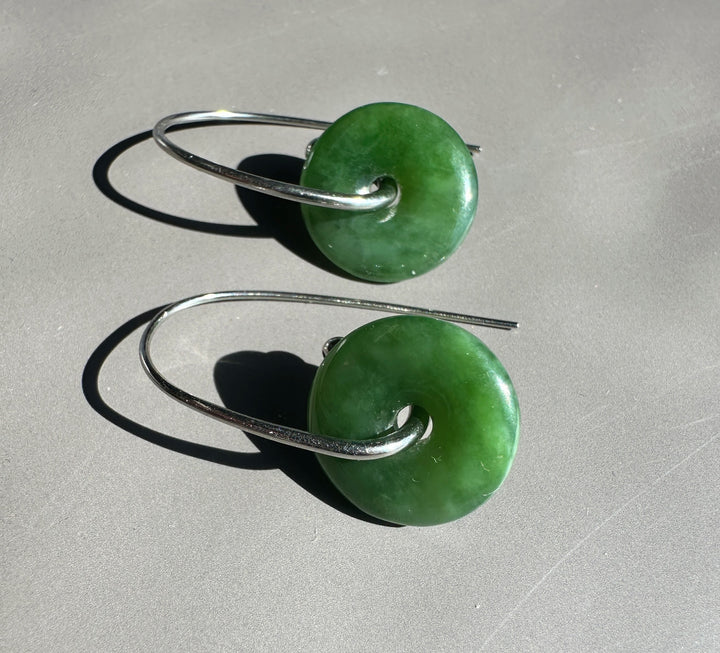 A pair of green stone donut earrings with silver wire hooks, displayed on a neutral gray surface with sharp shadows.