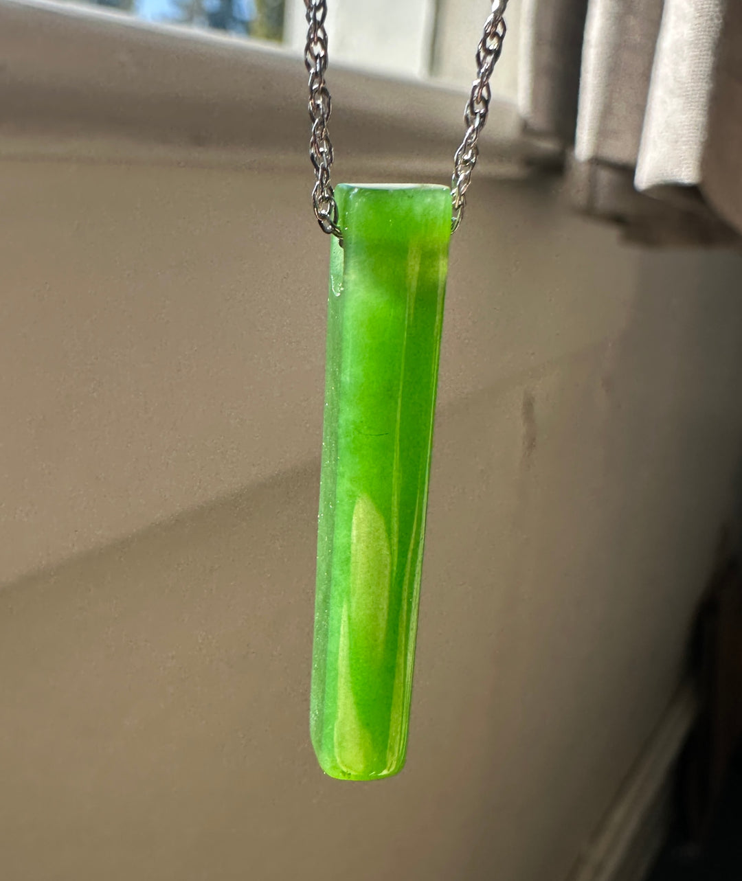 A bright green translucent rectangular pendant necklace hanging from a silver chain, photographed indoors against a beige wall near a window.