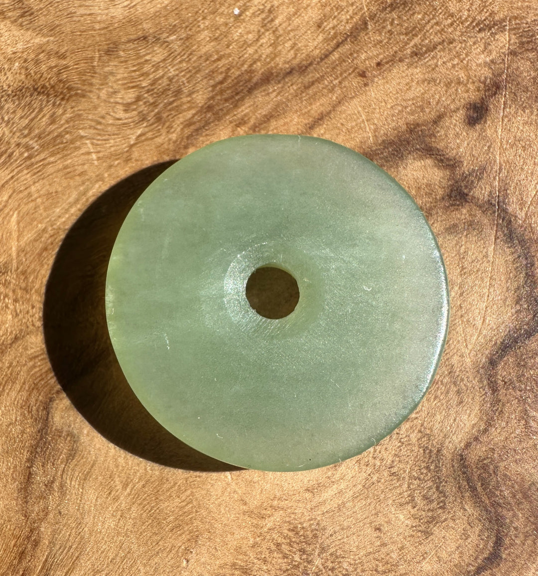 A round light green jade pendant with a central hole, resting on a textured wooden surface.