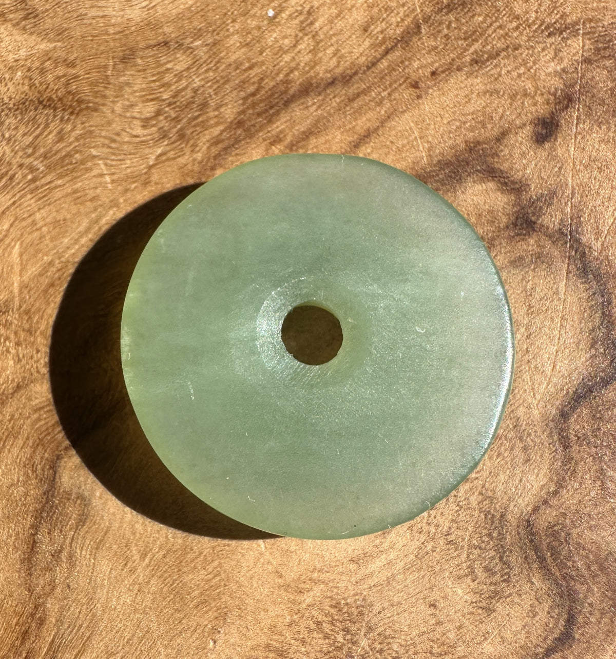 A round light green jade pendant with a central hole, resting on a textured wooden surface.