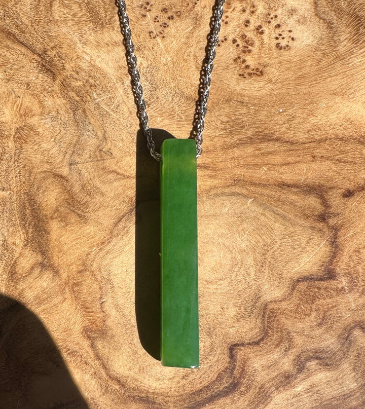 A green polished jade pendant necklace on a silver chain, resting on a textured wooden surface.