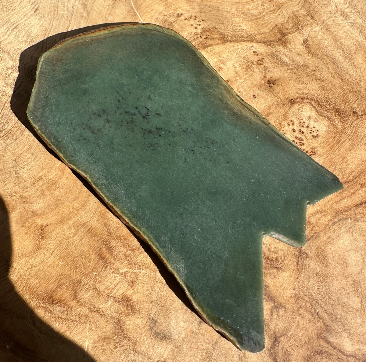 A dark green jade stone) with a natural irregular shape and rough outer edge, displayed on a textured wavy wood background.