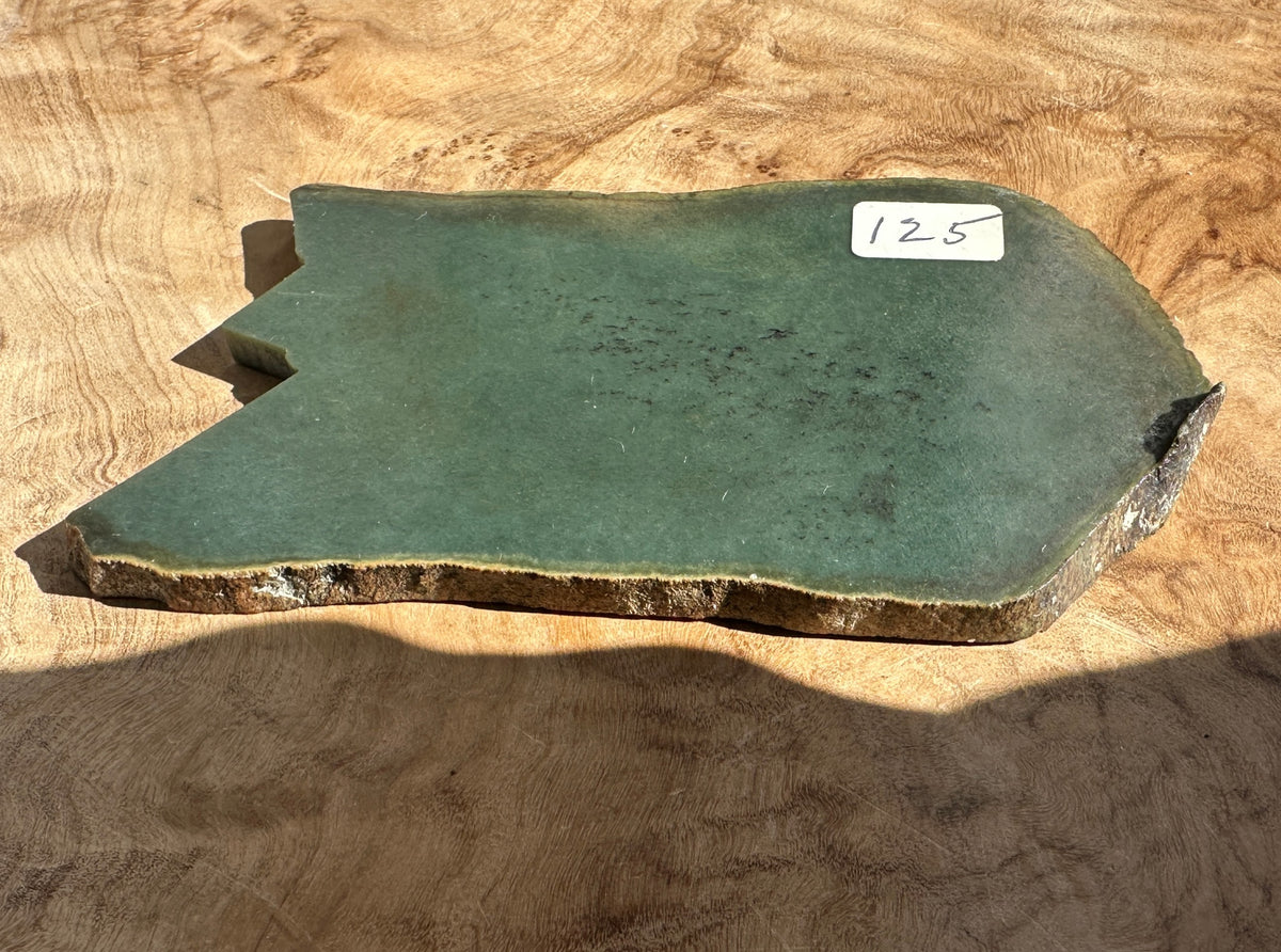 A dark green jade stone) with a natural irregular shape and rough outer edge, displayed on a textured wavy wood background.