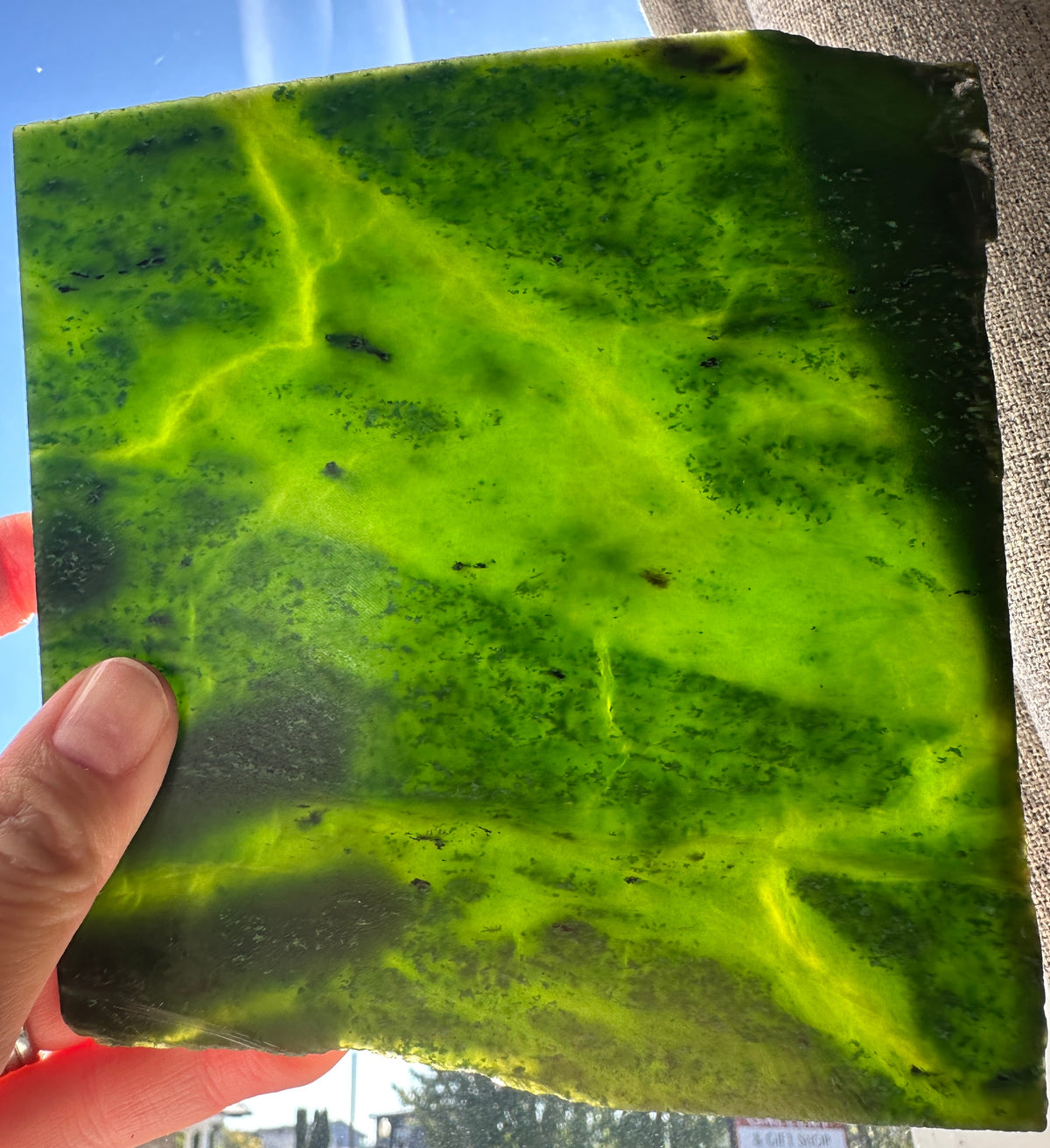 A hand holding a large, raw green stone slab with natural veining and dark inclusions, photographed in natural daylight.