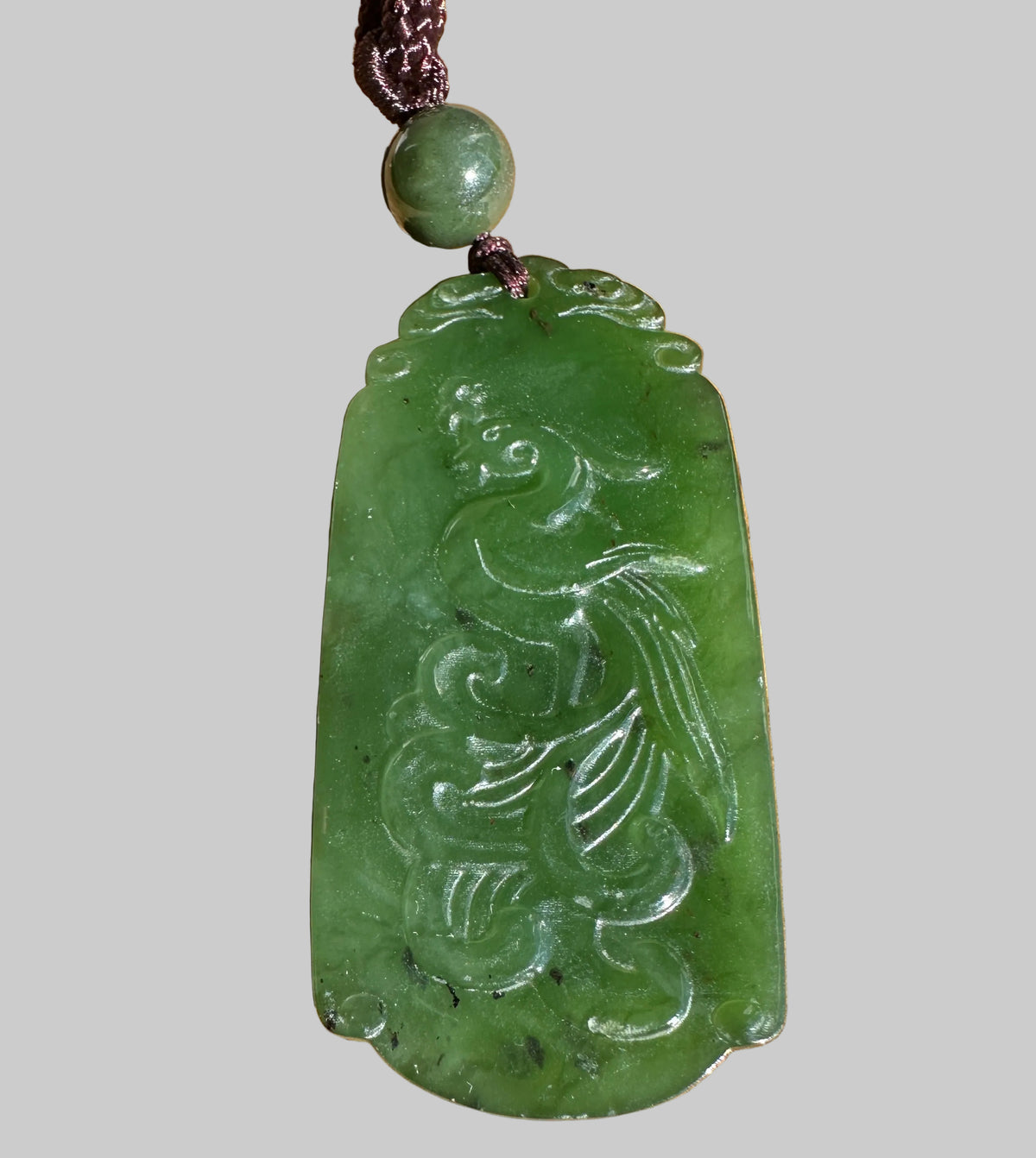 A green carved jade pendant featuring a phoenix design, hanging from a dark brown cord with a round green bead, set against a grey background.
