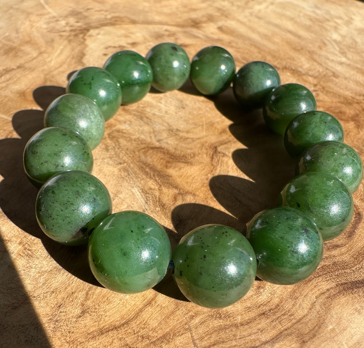 A beaded bracelet made of polished green stone beads with dark inclusions, displayed on a textured wooden surface.