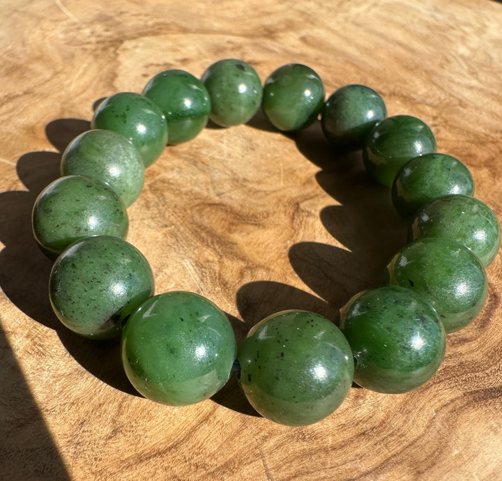 A beaded bracelet made of polished green stone beads with dark inclusions, displayed on a textured wooden surface.