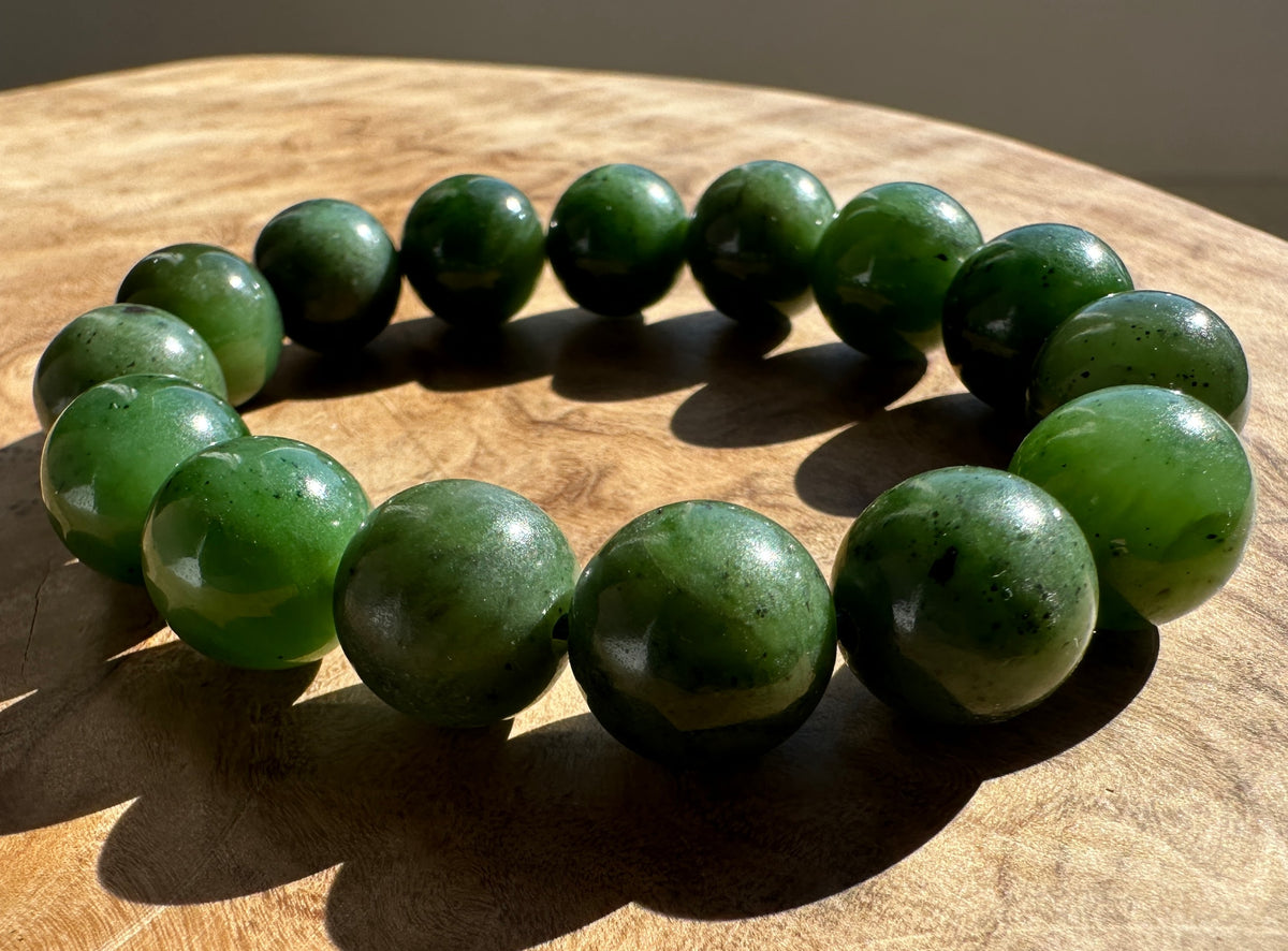 A beaded bracelet made of polished dark green jade-like round beads, displayed on a wooden surface in direct sunlight.