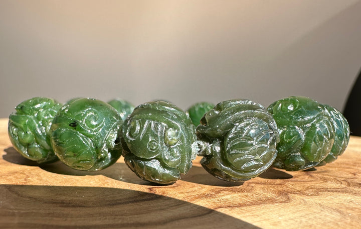 A beaded bracelet made of carved dark green jade-like round beads, displayed on a textured wooden surface.