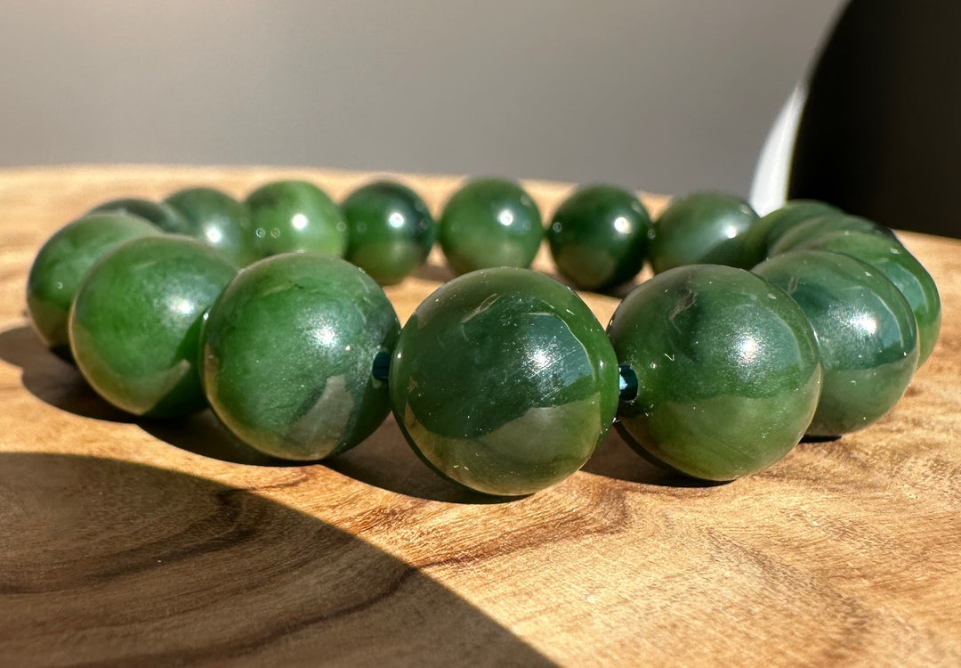 A beaded bracelet made of polished dark green jade-like round beads, displayed on a wooden surface with natural sunlight and shadows.
