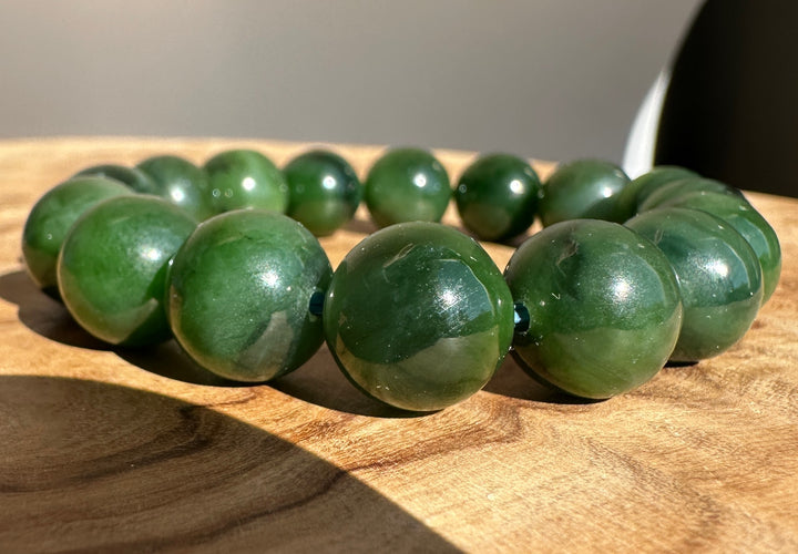 A beaded bracelet made of polished dark green jade-like round beads, displayed on a wooden surface with natural sunlight and shadows.