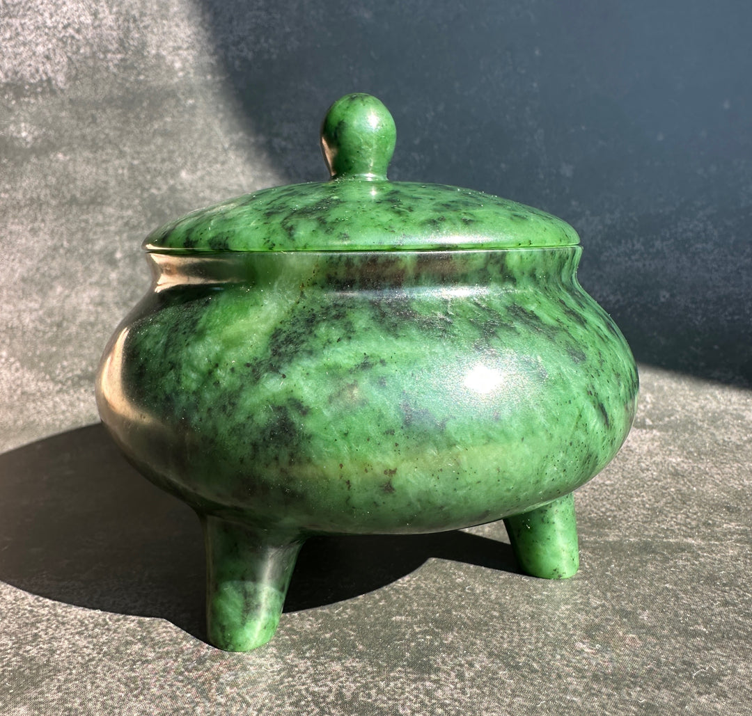 A polished green jade bowl with its matching lid placed beside it on a textured gray surface, photographed in bright natural sunlight.