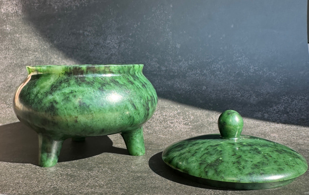 A polished green jade bowl with its matching lid placed beside it on a textured gray surface, photographed in bright natural sunlight.