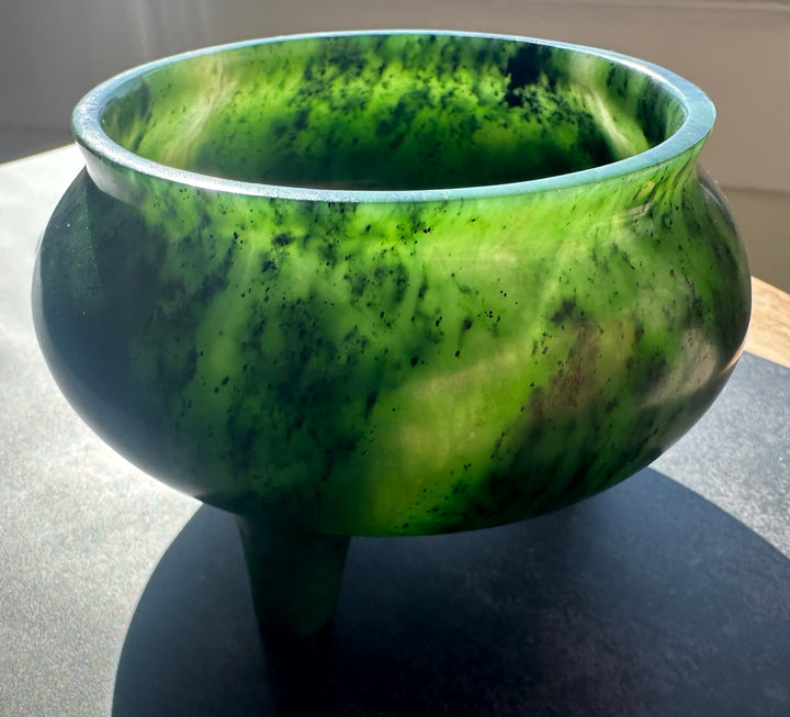 A green jade bowl with a three-legged tripod base, photographed on a dark surface with bright sunlight casting a shadow.