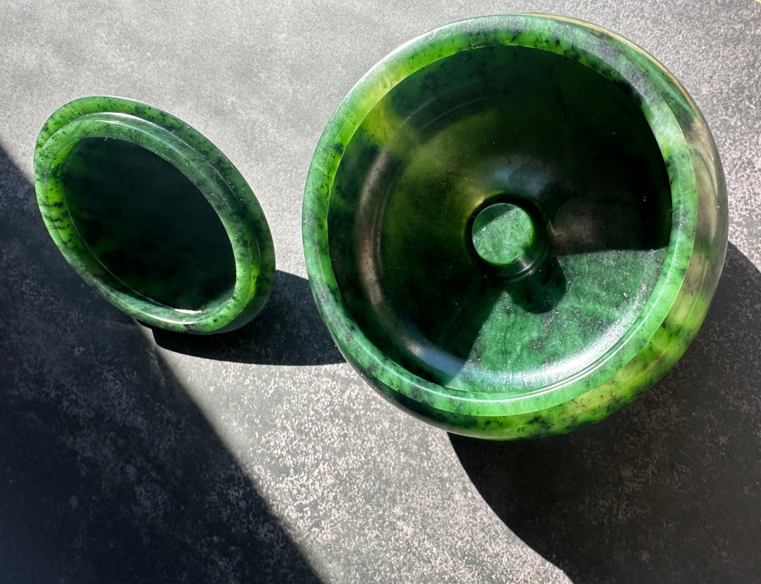 A polished green jade bowl with its matching lid placed beside it on a textured gray surface, photographed in bright natural sunlight.