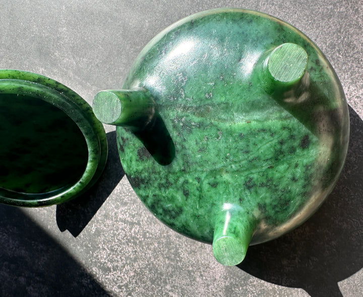 Bottom side of a polished green jade bowl with its matching lid placed beside it on a textured gray surface, photographed in bright natural sunlight.