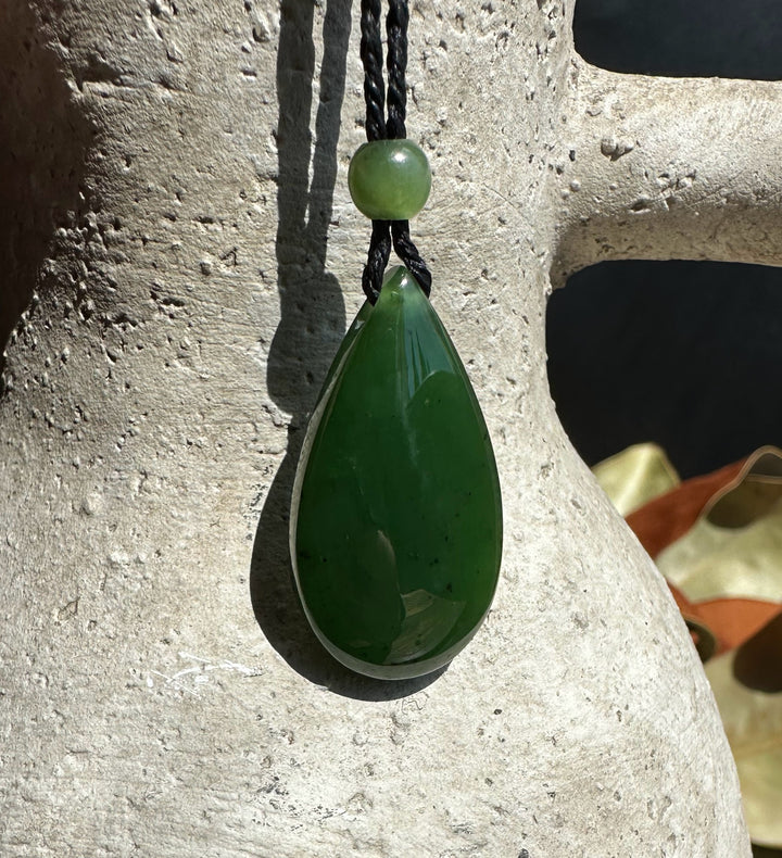 Green jade teardrop pendant with a round bead on a black cord against a textured stone background