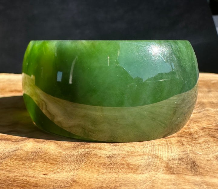 A polished green jade bangle with a glossy finish, resting on a natural wood surface against a dark background.