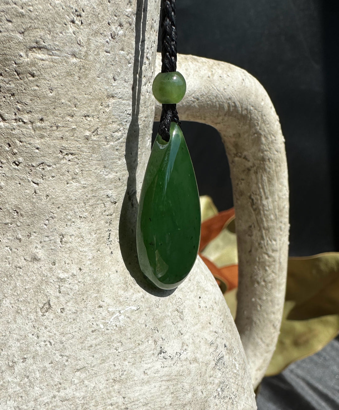 Jade teardrop pendant on a black braided cord with a small green bead against a textured stone background.