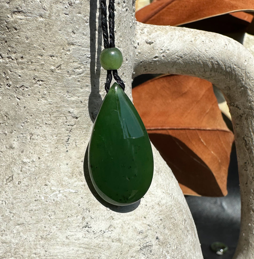Close up of jade teardrop pendant on braided black cord with a green bead, shown against a light stone surface.