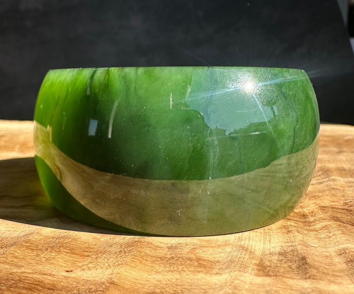A polished green jade bangle with a glossy finish, resting on a wooden surface against a dark background.