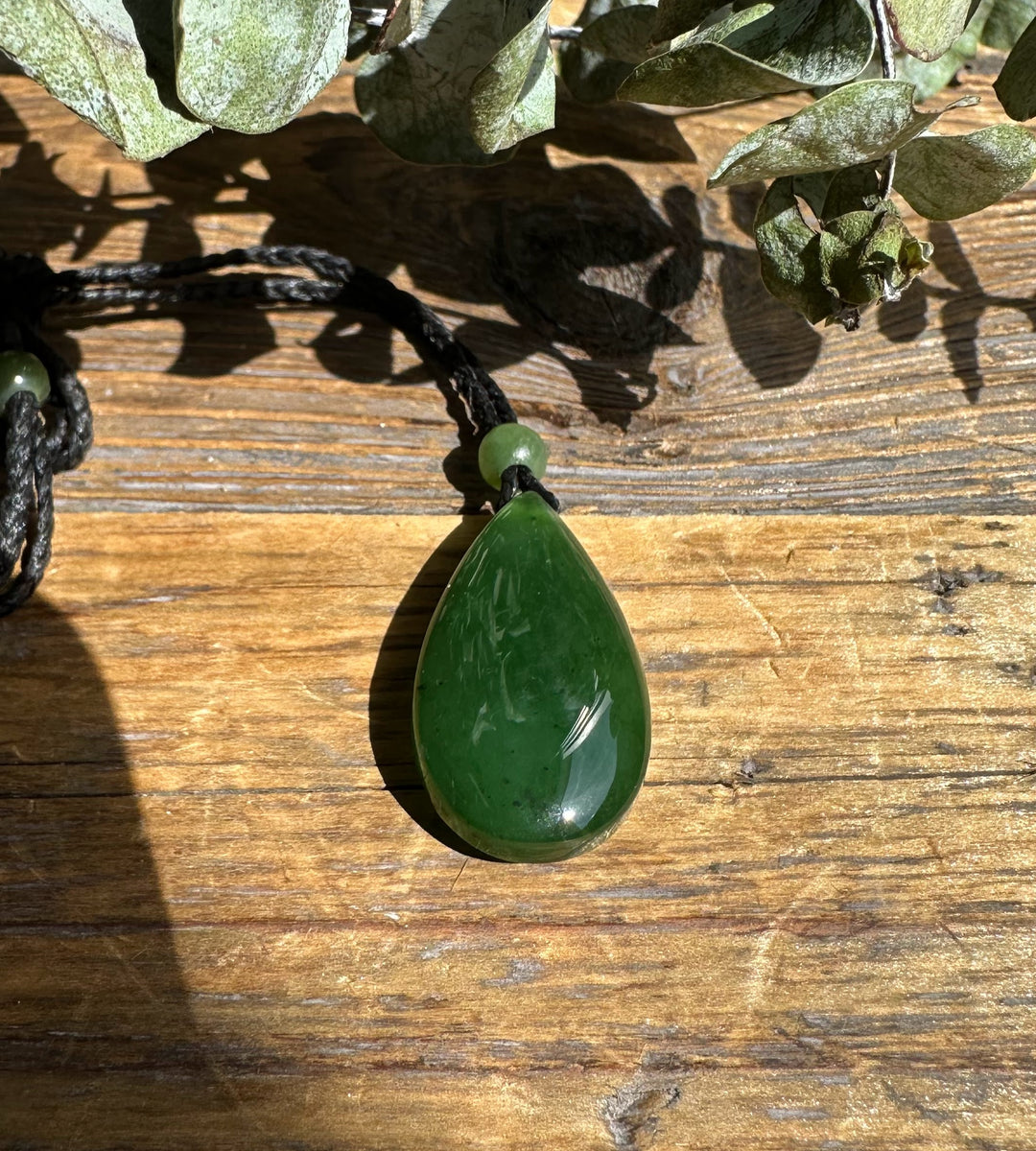 Jade teardrop pendant resting on a wooden surface with leaves in frame and natural sunlight.