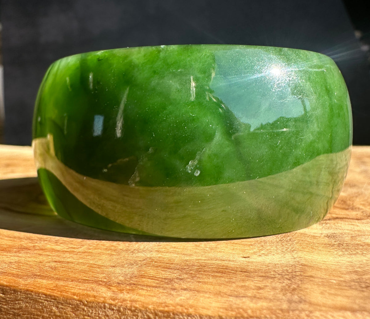A polished green jade bangle bracelet resting on a wooden surface with a dark background.
