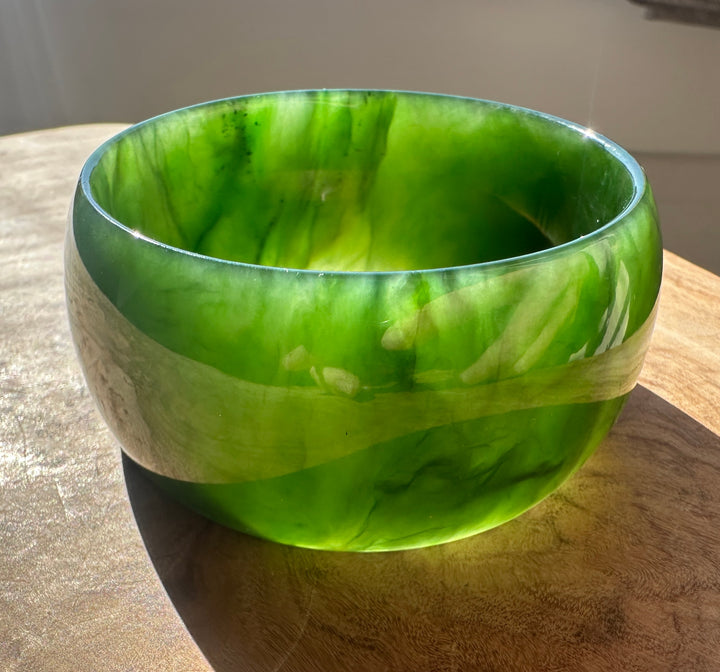 A polished green jade bangle with a glossy finish, resting on a natural wood surface against a dark background.