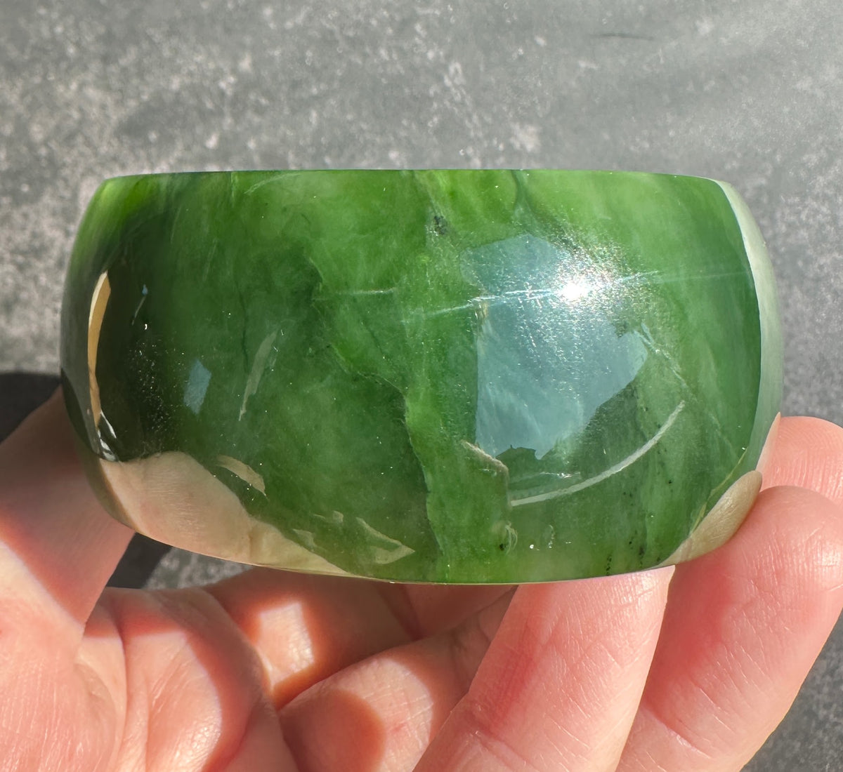 A polished green jade bangle bracelet held in a person's hand against a gray concrete background.