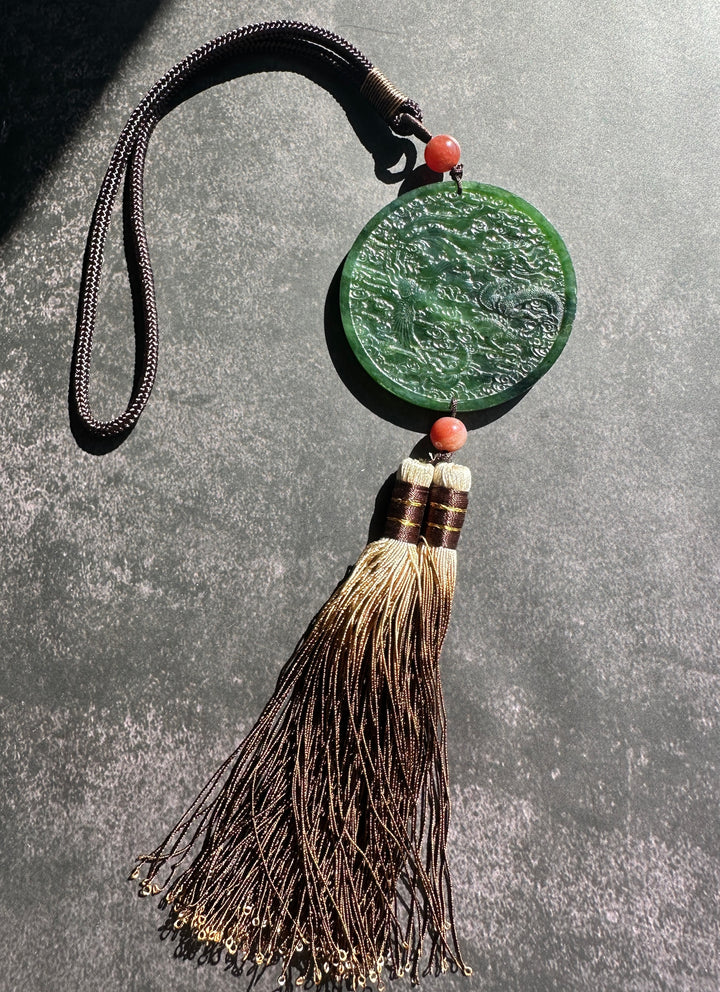 A green carved jade pendant with an embossed dragon design, hanging from a dark brown braided cord with a tassel and coral-colored beads, displayed on a textured gray background.