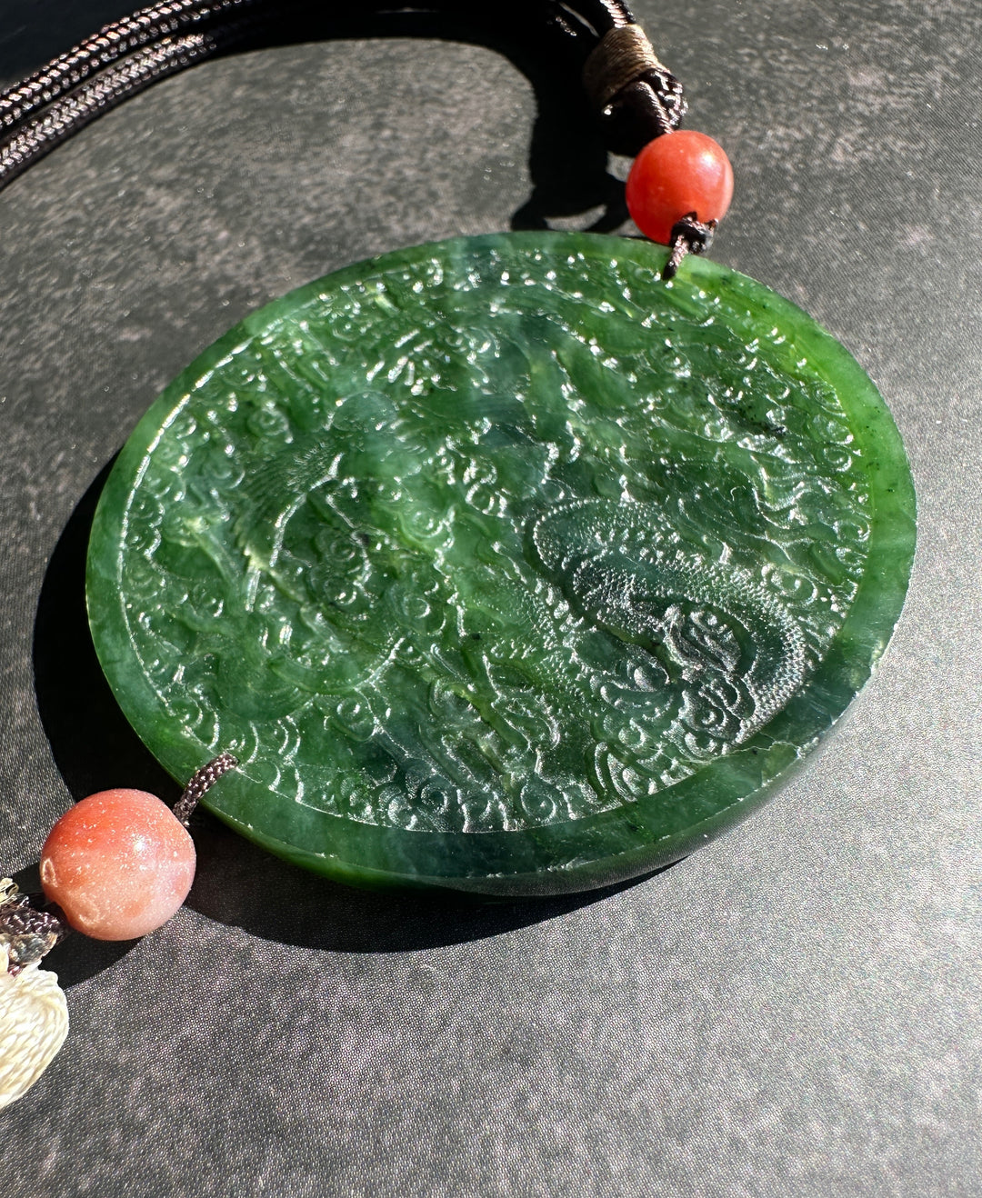 A round green jade pendant with carved dragon details, hanging from a dark cord with coral-red beads on a dark gray textured background.