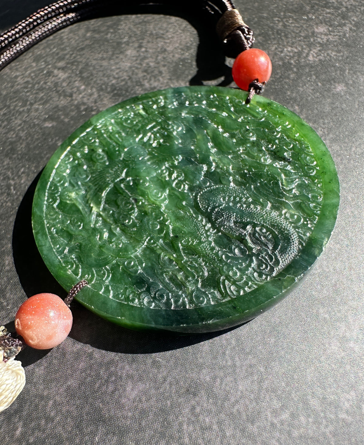 A round green jade pendant with carved dragon details, hanging from a dark cord with coral-red beads on a dark gray textured background.
