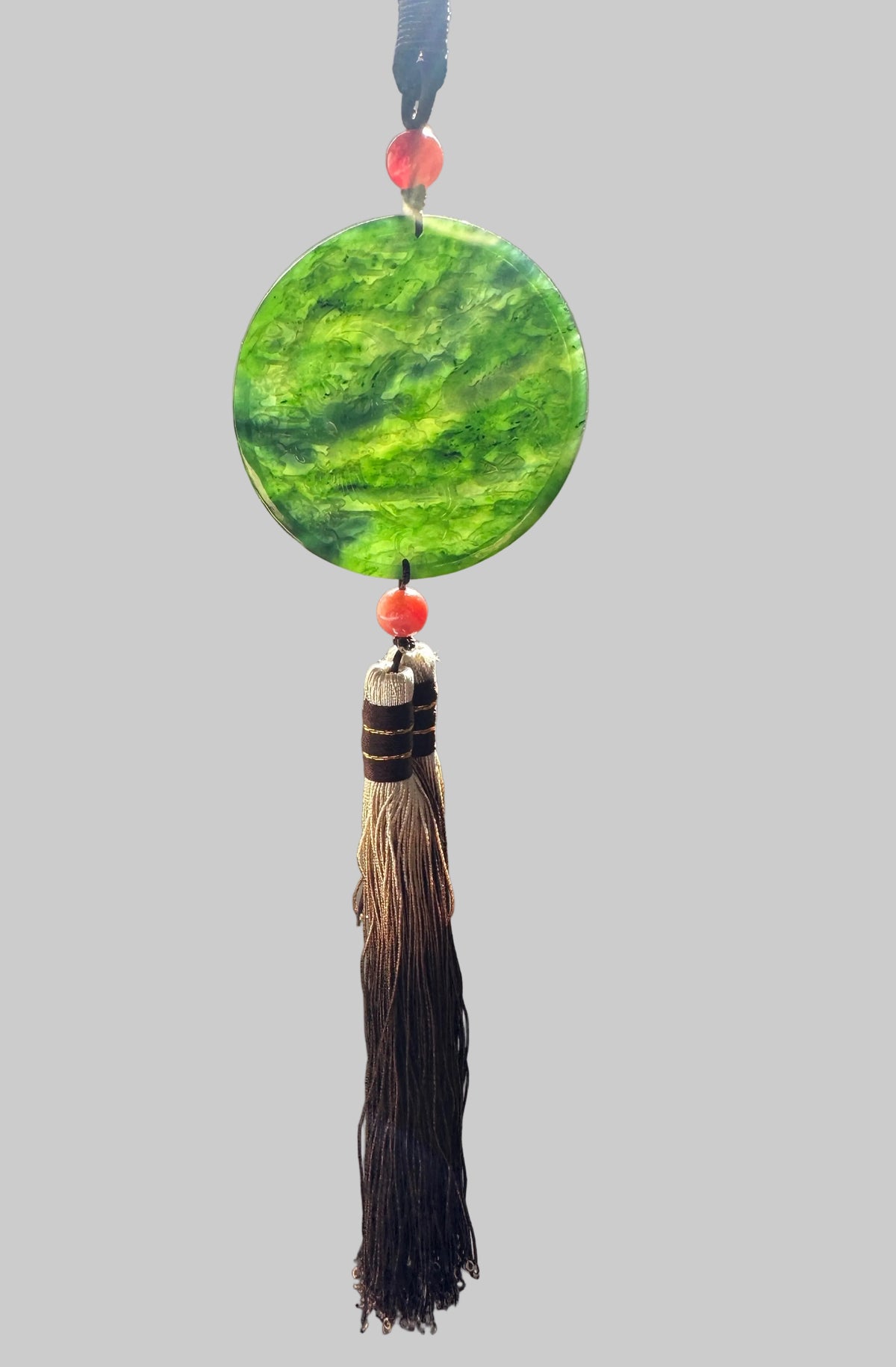 A circular green jade ornament with a brown tassel and red beads, hanging in front of a beige wall.