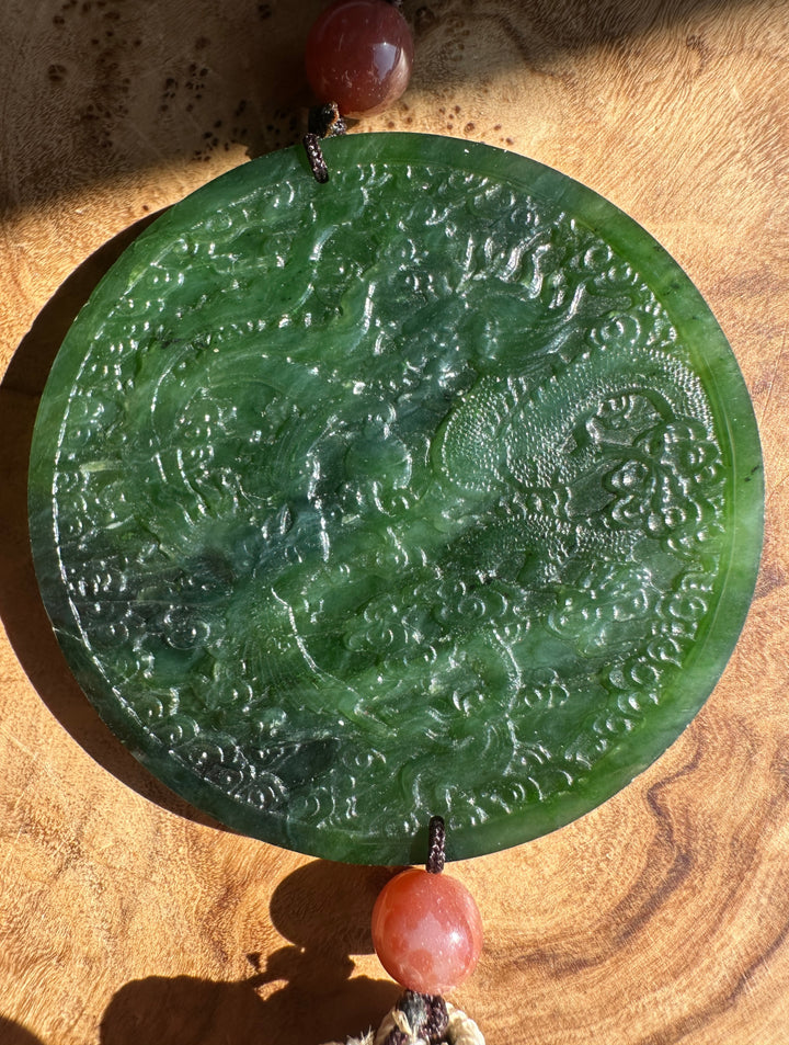 A round carved green jade pendant with dragon engravings, suspended from a cord with reddish-brown beads, displayed on a wooden surface.