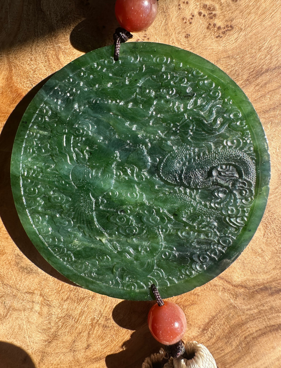 A round green jade pendant with an embossed dragon design, hanging from coral-red beads on a wooden surface.