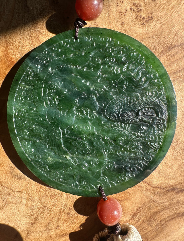 A round green jade pendant with an embossed dragon design, hanging from coral-red beads on a wooden surface.