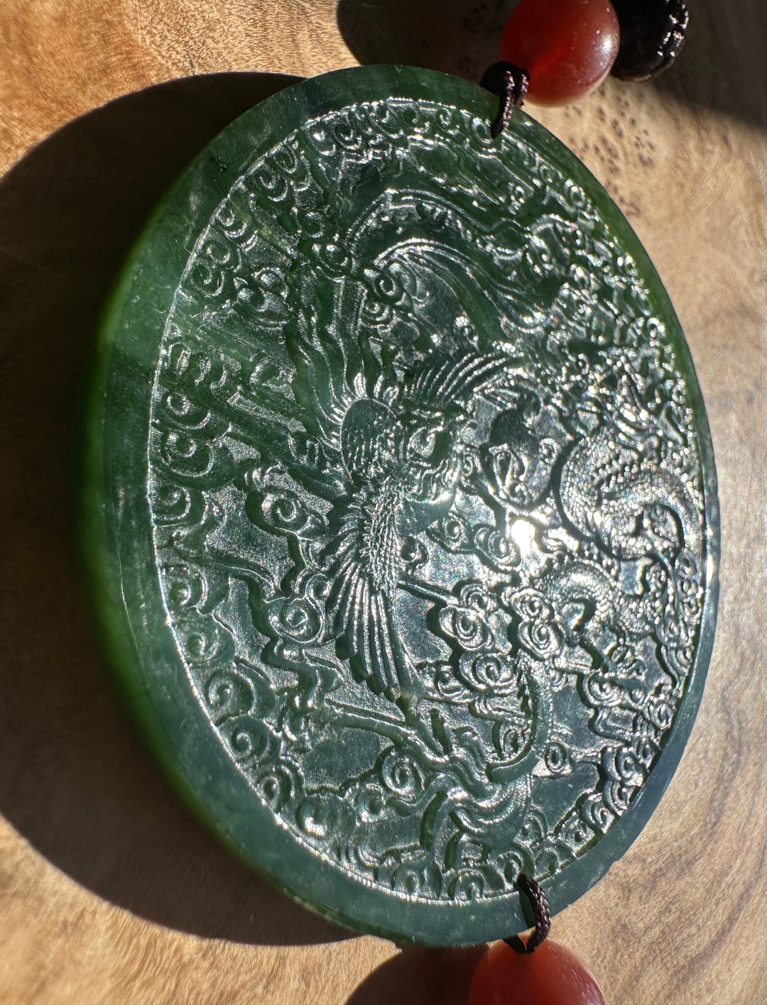A round carved jade pendant featuring a phoenix and cloud motif, displayed on a wooden surface with red beads at the top and bottom.