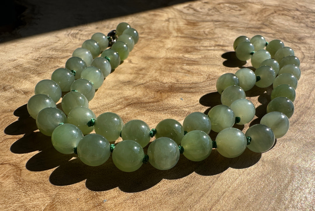 A strand of light green jade round beads with small green spacer accents and a silver clasp, laid out on a wooden surface in direct sunlight.