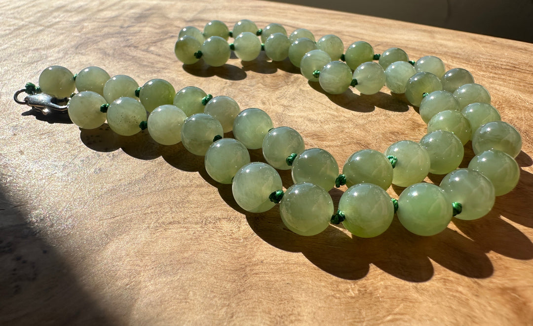 A strand of light green jade round beads with small green spacer accents and a silver clasp, laid out on a wooden surface in direct sunlight.