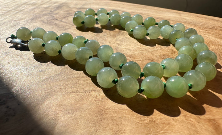 A strand of light green jade round beads with small green spacer accents and a silver clasp, laid out on a wooden surface in direct sunlight.