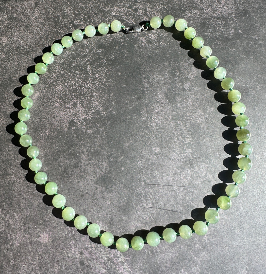 A beaded necklace made of round, polished light green gemstone beads with a silver clasp, displayed on a textured dark gray surface.
