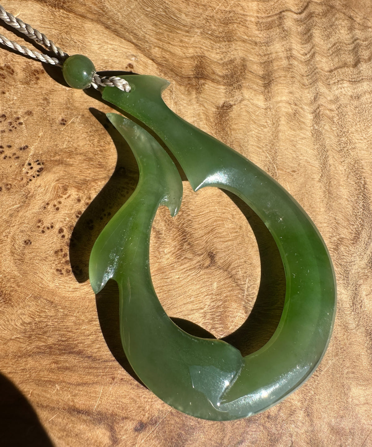 A polished green jade pendant necklace with a curved, hook-like shape, resting on a textured wooden surface.