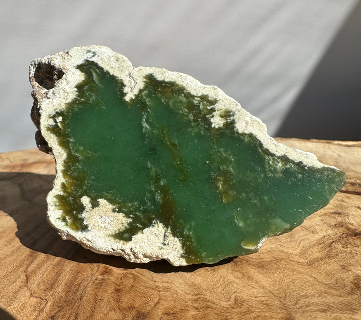 A raw green jade with a rough white crystalline border, displayed on a wooden surface with a neutral gray background.