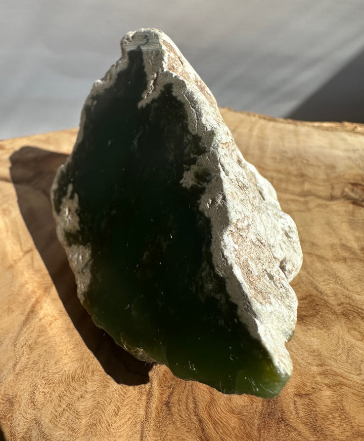 A raw green stone or mineral specimen with a rough, textured light gray outer surface, standing upright on a wooden surface.
