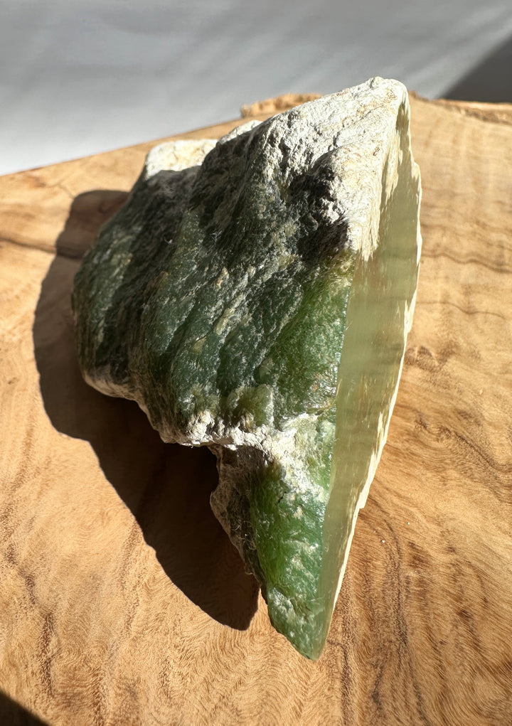 A raw green stone or mineral specimen with rough outer texture and a polished green inner face, displayed on a wooden board in natural light.