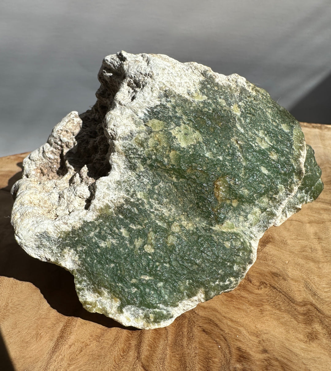 A raw green stone or mineral specimen with a rough white and gray crystalline outer surface, displayed on a curved wooden surface against a neutral gray background.