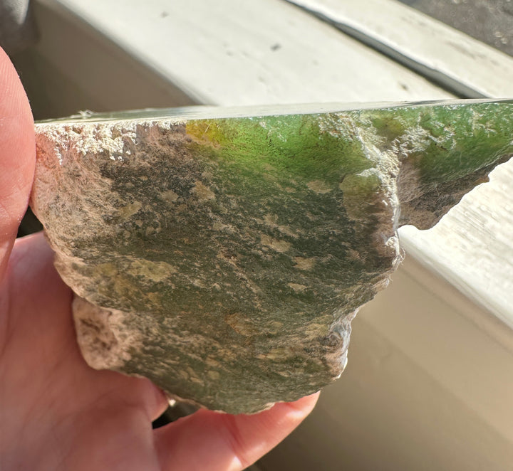 A hand holding a rough, irregularly shaped piece of green translucent material, likely a raw gemstone or crystal, with a textured, earthy base and a smooth green top surface.