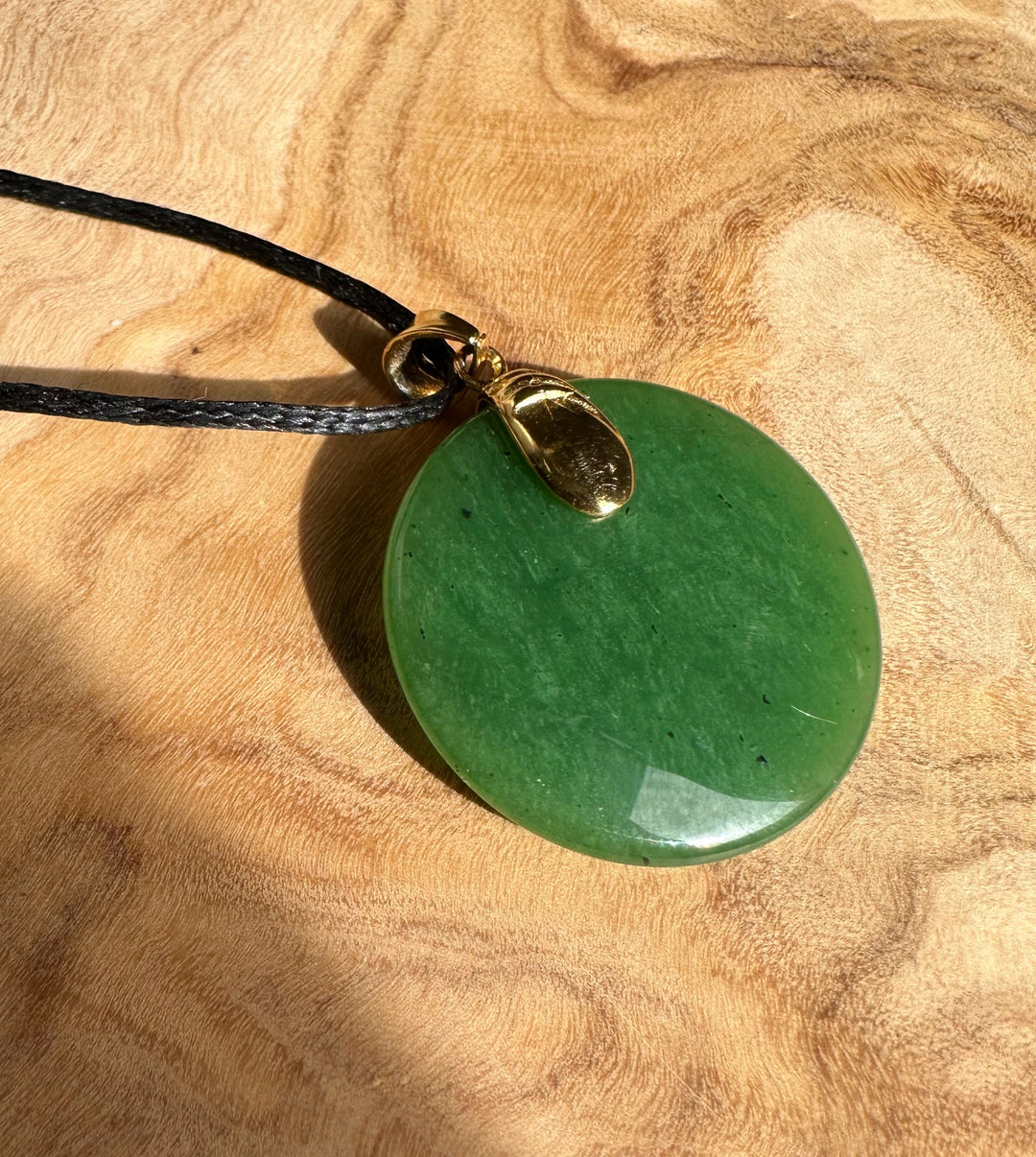 Round green jade pendant with gold bail on black cord resting on wood