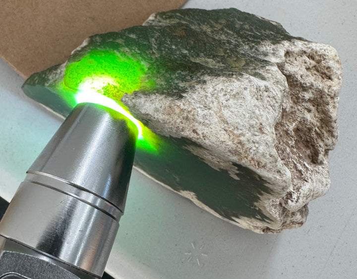 A close-up of a rough green stone or mineral slab with a bright green glow being projected onto it by a handheld metal light source. The stone has a natural rough texture with dark green, gray, and off-white areas.