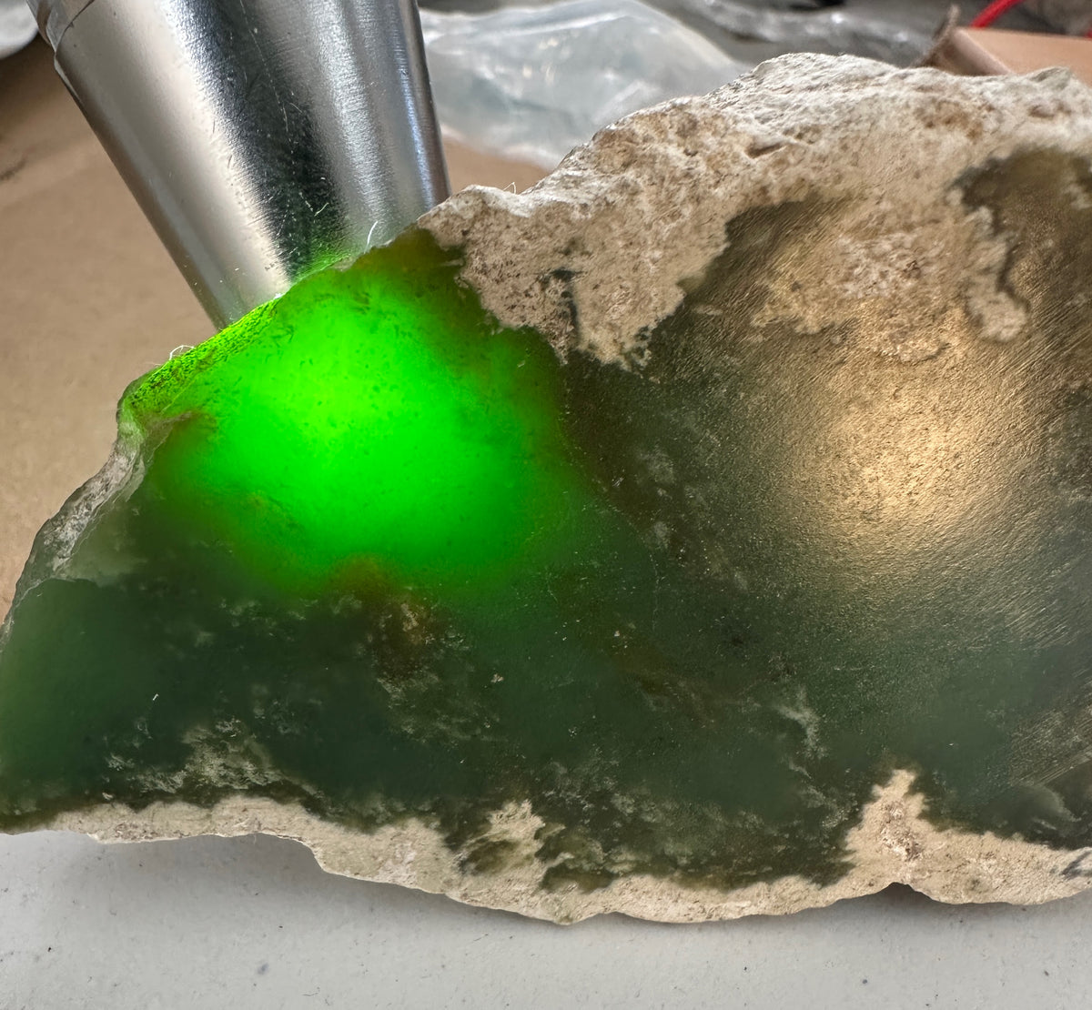 A raw green stone or gemstone slab with rough edges, showing a bright fluorescent green area under light, with a metallic light source visible at the top left.